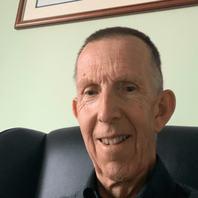 Dave is sat on a black armchair. He is smiling at the camera. He has short dark hair and is wearing a shirt with a collar. The wall behind him is light green and there's a framed picture of a train on the wall behind him.
