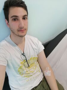 Ziyad is sitting down and taking a selfie. He’s wearing a white shirt with glasses hanging from the centre. A white dressing is taped over the inside of his elbow where bloods have been taken. He looks tired. 