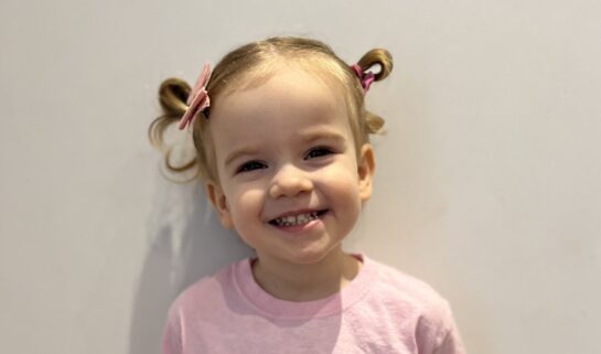 Imogen is smiling at the camera with her ponytails tied into playful loops that stick up in the air. She’s wearing a pink shirt that reads: “Coeliac PRINCESS” with a tiara above the words.