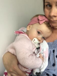 Kerry is looking at the camera while holding Imogen in her arms. Imogen is wearing a pink headband and using a dummy, dressed in a light pink cardigan.