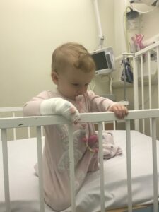 Imogen is kneeling and propping herself up against a white crib. She’s wearing a pink baby onesie, with a baby monitor visible in the background.