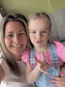 Kerry and Imogen are smiling at the camera. Imogen is making a pouty face and holding up a peace sign with her fingers. Kerry is wearing a white blouse, and Imogen is dressed in blue overalls with a pink shirt underneath. They are standing in front of a green wall.