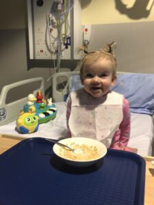 Imogen is sitting on a hospital bed with toys beside her. A bowl of cereal rests on her bed table. She’s smiling at the camera, wearing a food bib, with her ponytails tied up into little loops that stick up in the air.
