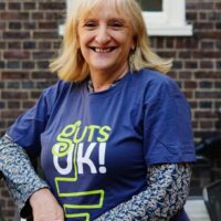 Julie has short blonde hair and is wearing a short sleeved blue t-shirt with Guts UK written in green and white. She has a long sleeved patterned top underneath. She is standing outside, in front of a building with brown bricks and white windows. Her arm is leaning on a railing.