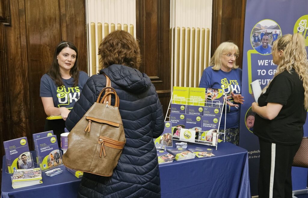 Two of our team members are stood behind an information stand, with Guts UK leaflets and posts. They are talking to two people of the other side of the table.