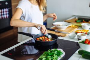 A person, who has long blonde hair and os wearing a white short sleeved t-shirt, is stood at a kitchen bench. In front of them is a hob with a pan on top. The pan has food in it, and they are using a kitchen utensil to move the food as it cooks. On the other side of the hob, there is a plate with salad leaves next to it. On the bench beside the hob, there is a wooden chopping board, a knife, and some bowls. 