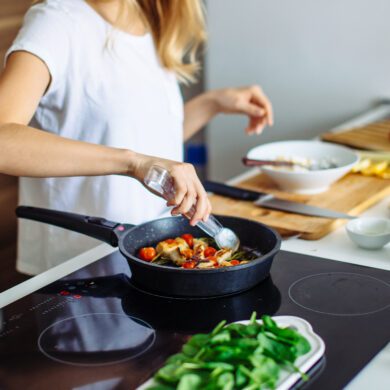 A person, who has long blonde hair and os wearing a white short sleeved t-shirt, is stood at a kitchen bench. In front of them is a hob with a pan on top. The pan has food in it, and they are using a kitchen utensil to move the food as it cooks. There is a plate with salad leaves next to it. On the bench beside the hob, there is a wooden chopping board, a knife, and a bowl.