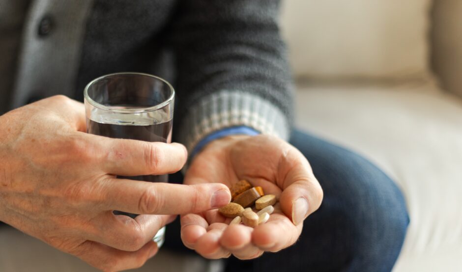 A man is sat down. He is holding a glass of water in one hand and some tablets in another. He is wearing blue jeans and a dark grey jumper.