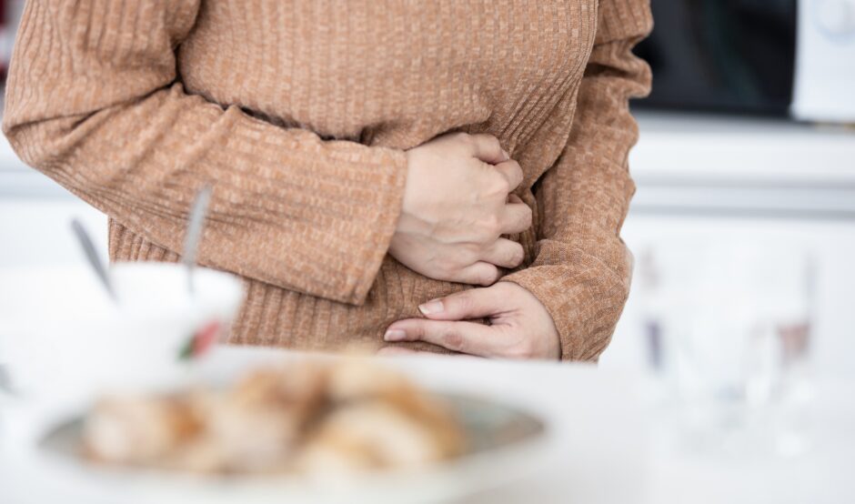 A person's torso is seen, covered by a light brown jumper. They arms are covering their stomach, with their hands gripping their upper abdomen, indicating pain or discomfort.