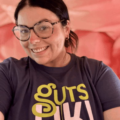 Jade, Guts UK's Senior Communications Manager, is smiling at the camera and wearing a blue Guts UK t-shirt. She has dark hair, tied up and is wearing round glasses with a dark frame.