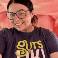 Jade, Guts UK's Senior Communications Manager, is smiling at the camera and wearing a blue Guts UK t-shirt. She has dark hair, tied up and is wearing round glasses with a dark frame.