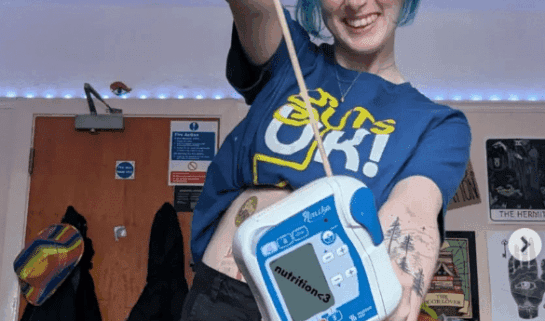 Luce is smiling at the camera, holding up her feeding pump. She wears a blue Guts UK t-shirt and black shorts and has light blue hair. She has temporary #GUTSelfie tattoos on her legs.