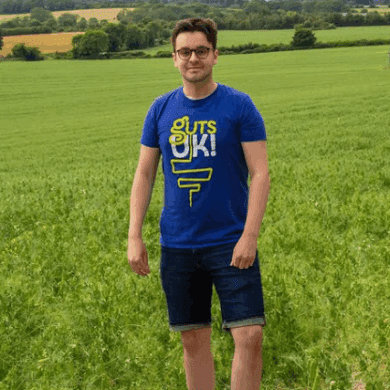 Daniel is wearing a Guts UK t-shirt and dark denim shorts, with black trainers. He wears black rimmed glasses and stands in a lush, green field, with electricity pylons down the middle. He is smiling.