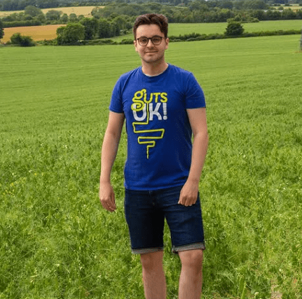 Daniel is wearing a Guts UK t-shirt and dark denim shorts, with black trainers. He wears black rimmed glasses and stands in a lush, green field, with electricity pylons down the middle. He is smiling.