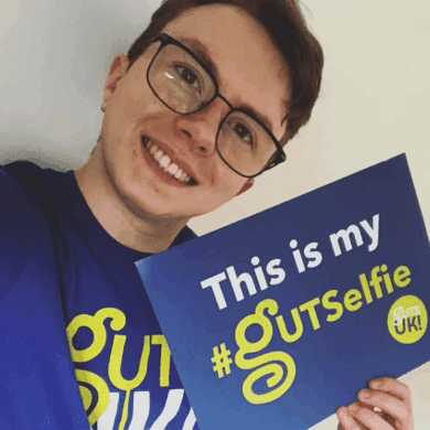 A #GUTSelfie of a boy wearing black, rectangular, curved frame glasses. He has short brown hair and is smiling. He wears a blue Guts UK t-shirt and holds a board up which reads 'This is my #GUTSelfie'.