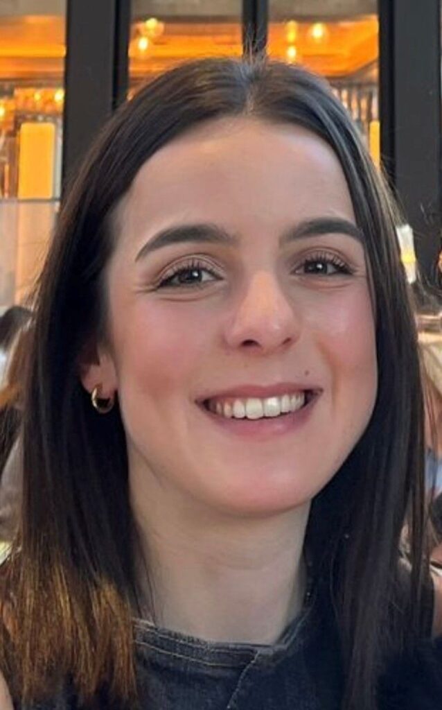 Katerina has long dark brown hair and is smiling at the camera in this headshot
