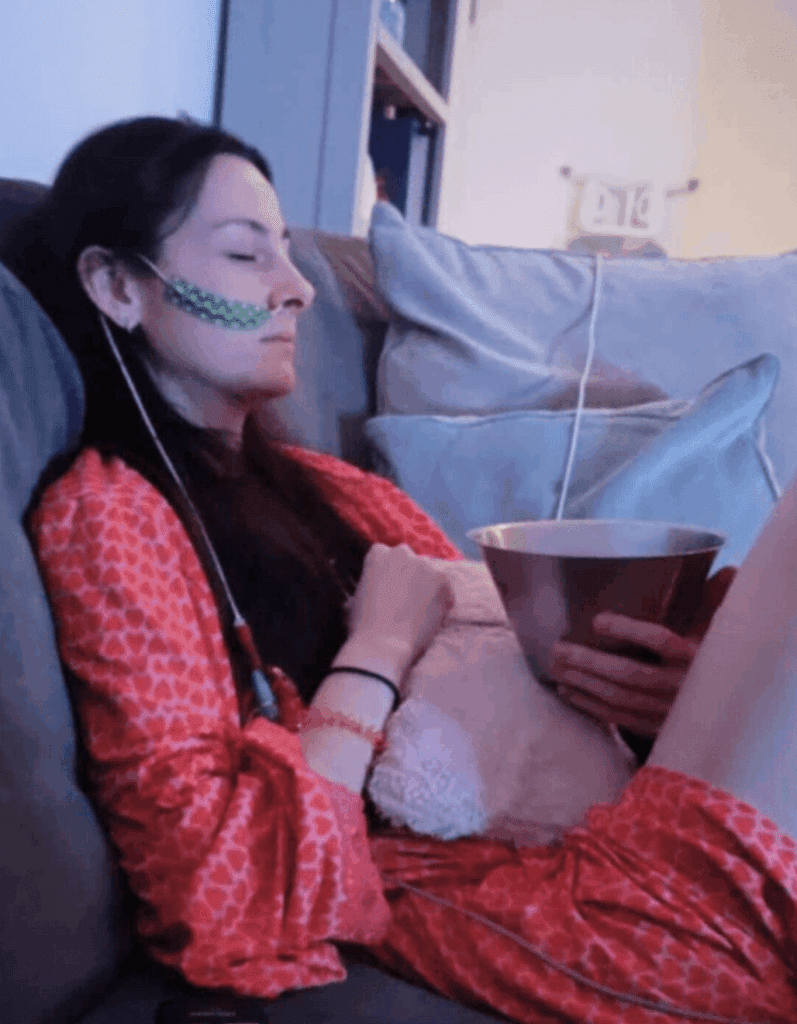 Jessie is sat on her sofa, wearing a pink dressing gown with red hearts all over it. She has her eyes closed, is holding a silver sick bowl and has a light pink hot water bottle on her knee. She has a feeding tube going into her nose, with green and white polka dot tape securing it to her cheek.