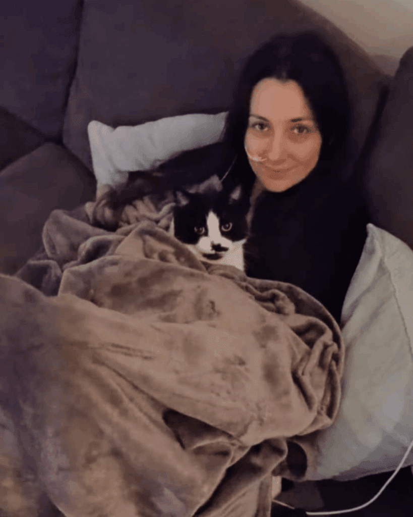 Jessie is on her sofa, cuddled under a medium brown fleece blanket with one of her cats. She wears a black jumper and has a feeding tube going into her nose.