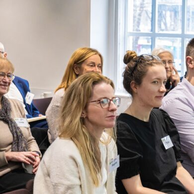 A group of people sit together in an audience, listening to a speaker.