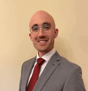 Dr Thomas Conley smiles at the camera. He is wearing a white shirt, a red tie and a grey suit jacket.
