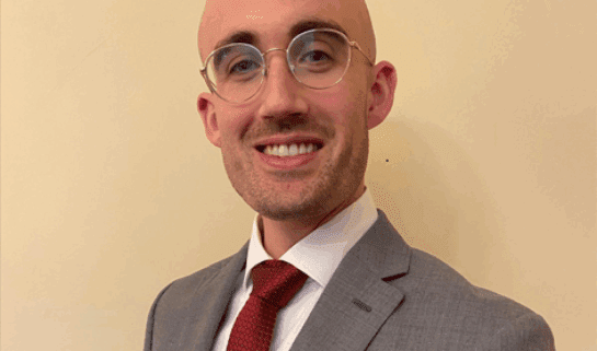 Dr Thomas Conley smiles at the camera. He is wearing a white shirt, a red tie and a grey suit jacket.