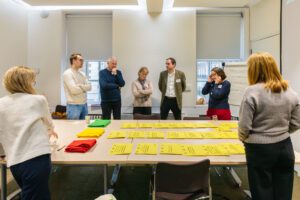 A group of people stand around a table, talking to one another. There is a table in the centre of the room, with pieces of paper set out in front of green, yellow and red pieces of material.