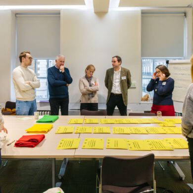 A group of people stand around a table, talking to one another. There is a table in the centre of the room, with pieces of paper set out in front of green, yellow and red pieces of material.