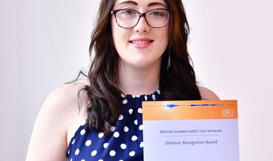 Chloe smiles at the camera, holding her certificate. She has black rimmed, rectangular, curved glasses and is wearing a navy dress with white polka dots on. She has brown hair, just below shoulder length.