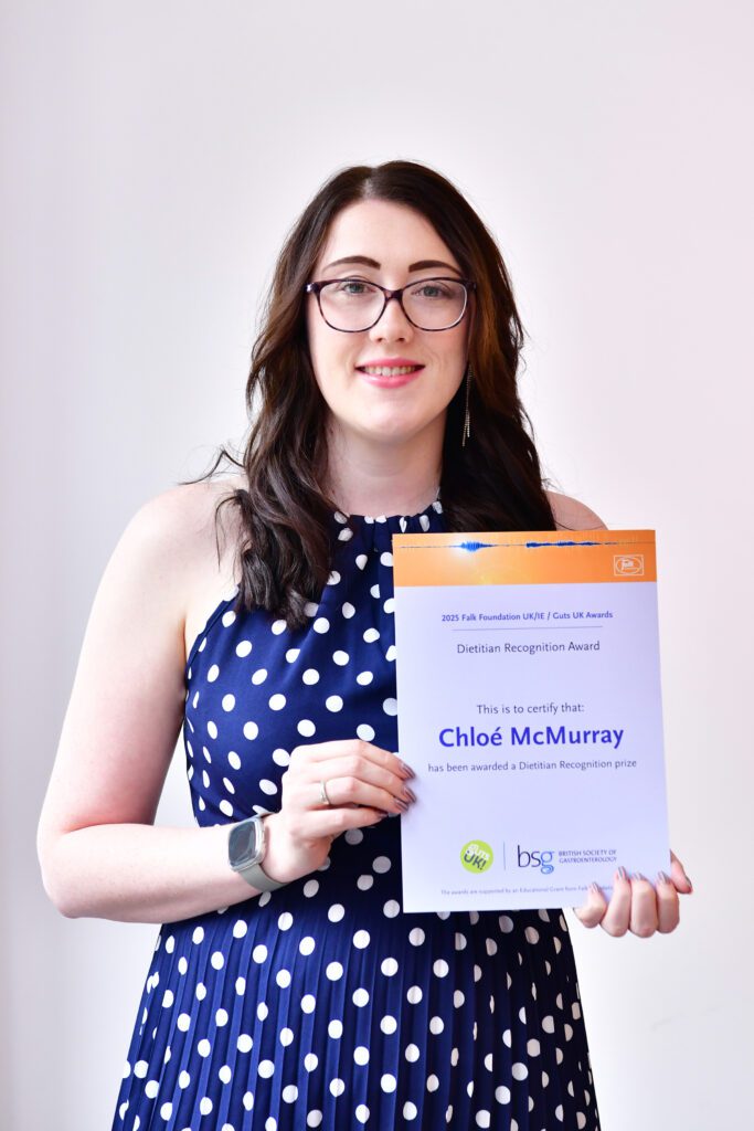 Chloe smiles at the camera, holding her certificate. She has black rimmed, rectangular, curved glasses and is wearing a navy dress with white polka dots on. She has brown hair, just below shoulder length.