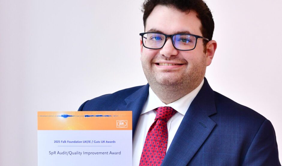 Munir smiles at the camera, holding his certificate. He has short black hair, is wearing black rimmed glasses and wears a navy suit, with a white shirt and red tie underneath.