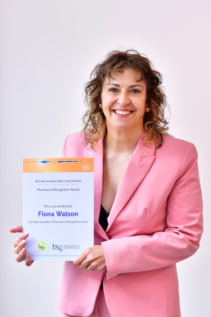 Fiona smiles at the camera, holding her certificate. She has shoulder length, curly, brown hair and wears a light pink blazer.