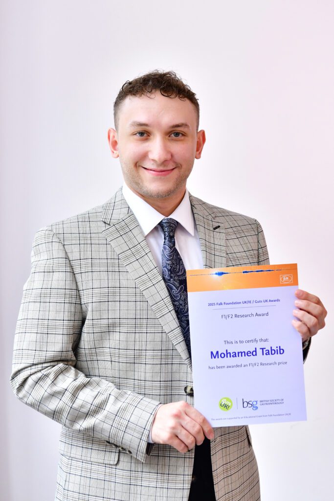 Mohamed smiles at the camera, holding his certificate. He wears a light grey, plaid suit and has short, light brown hair.