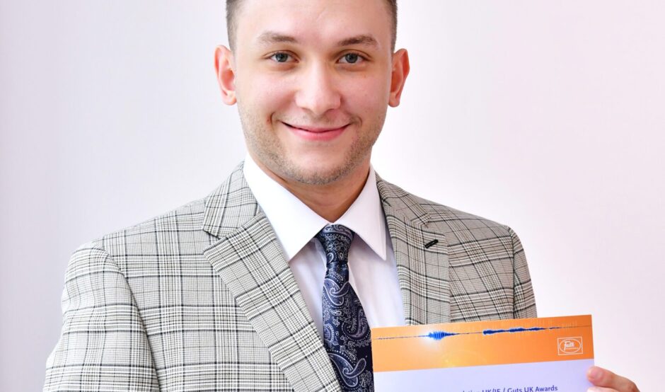 Mohamed smiles at the camera, holding his certificate. He wears a light grey, plaid suit and has short, light brown hair.