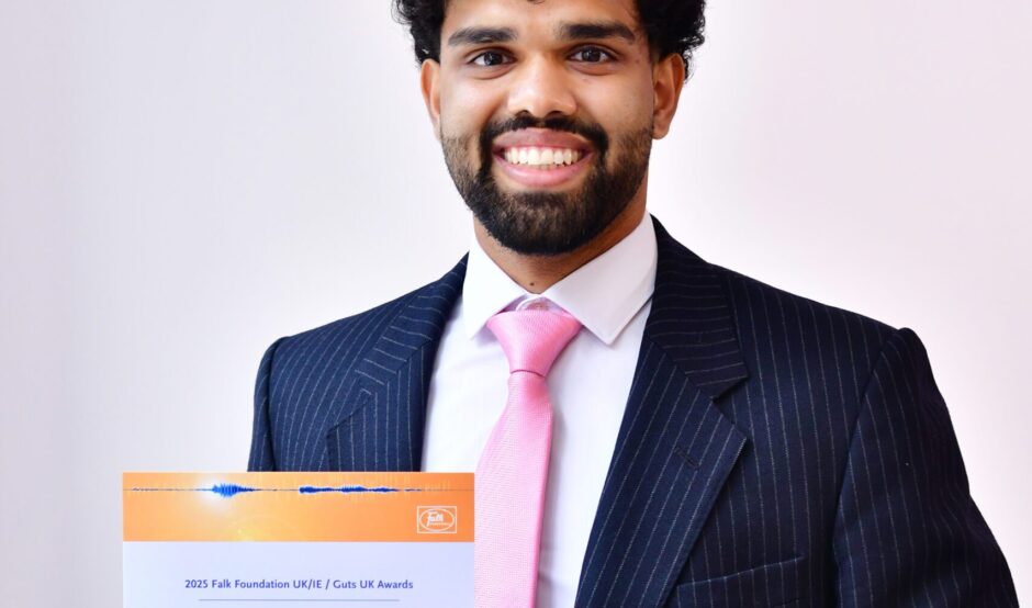 Nikhil smiles at the camera, holding his certificate. He wears a deep navy suit, a white shirt and light pink tie. He has black hair.