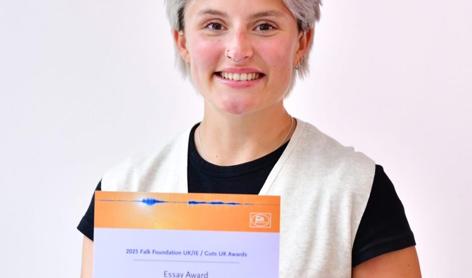 Nat smiles at the camera, holding her certificate. She has short, blonde hair and wears a black t-shirt, with a white vest cardigan over the top, and grey trousers.