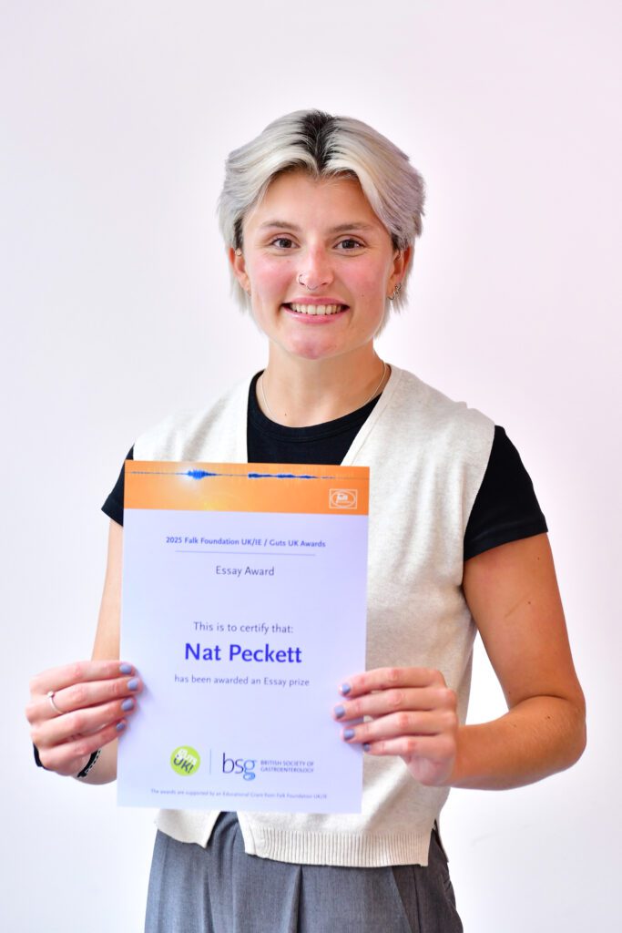 Nat smiles at the camera, holding her certificate. She has short, blonde hair and wears a black t-shirt, with a white vest cardigan over the top, and grey trousers.