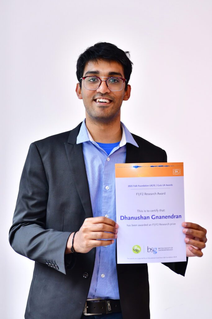 Dhanushan is standing smiling with his Awards certificate. He wears a light blue shirt and grey suit.