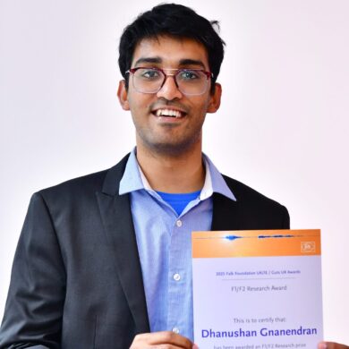 Dhanushan is standing smiling with his Awards certificate. He wears a light blue shirt and grey suit.