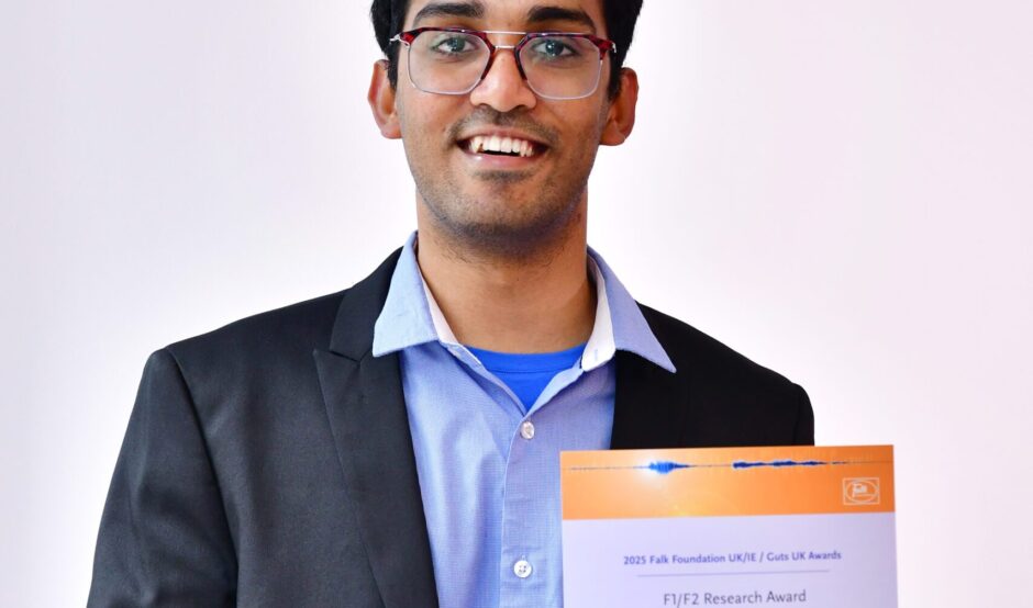 Dhanushan is standing smiling with his Awards certificate. He wears a light blue shirt and grey suit.