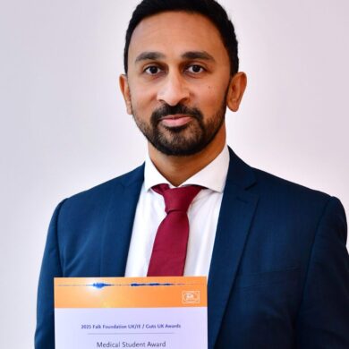 Amar smiles at the camera, holding his certificate. He has short, black hair, wears a navy suit, white shirt and deep red tie.