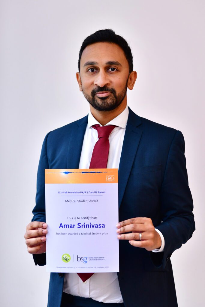 Amar smiles at the camera, holding his certificate. He has short, black hair, wears a navy suit, white shirt and deep red tie.