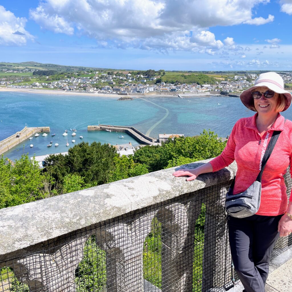 Linda is in the foreground of the picture. She has a short, blonde bob and wears black sunglasses, and a light pink sunhat. She wears a bright pink fleece three-quarter zip jumper, and navy trousers with a grey cross-body bag. In the background, there is a harbour with bright blue water, and white boats dotted around. Further behind them, there is a shoreline and houses in the distance, surrounded by lush green scenery, and bright blue skies with fluffy, white clouds.