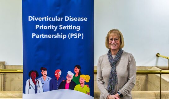 Linda has short, straight, blonde hair styled in a bob, wears black rimmed glasses and wears a beige jumper, blue floral scarf and black trousers. She smiles at the camera, stood next to a narrow roller banner with a Guts UK navy background which has white text on it, reading 