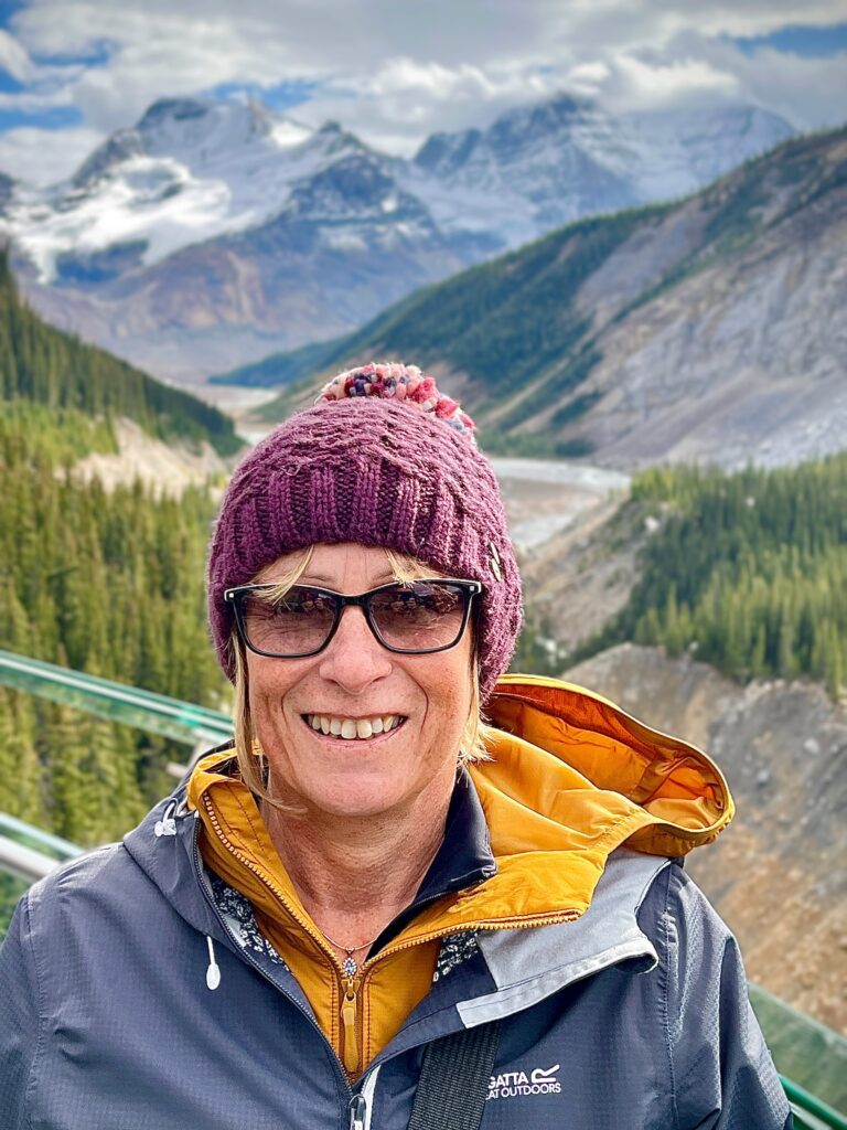 A close-up of Linda smiling at the camera. She smiles, and has short, blonde hair in a bob and wears black sunglasses. She wears a burgundy bobble hat, and has a yellow undercoat on, with a navy Regatta waterproof on top. There are rolling mountains in the background and forest greenery, partially covered by white clouds.