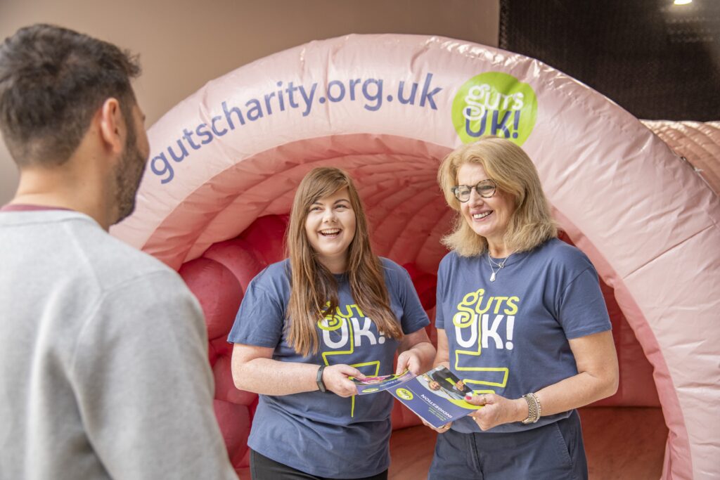 Two members of the Guts UK team - Diane and Helen - stand in front of Colin our inflatable colon. They wear Guts UK branded t-shirts and hold our patient information leaflets in their hands. They are smiling at a man, who has his back to the camera. He has short dark hair and wears a long sleeved grey sweatshirt.