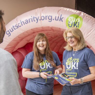 Two members of the Guts UK team - Diane and Helen - stand in front of Colin our inflatable colon. They wear Guts UK branded t-shirts and hold our patient information leaflets in their hands. They are smiling at a man, who has his back to the camera. He has short dark hair and wears a long sleeved grey sweatshirt.