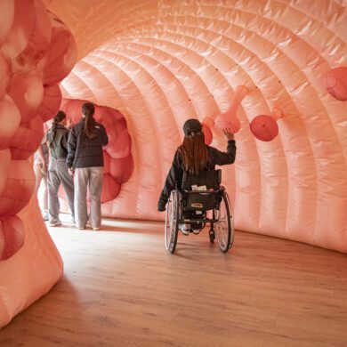 Three people take a tour through Colin the inflatable colon. One person is sat in a wheelchair and has their arm reaching out to touch one of the polyps on the wall of the colon. Further into the colon, two people are stood up colon looking at the bowel cancer on the colon wall.
