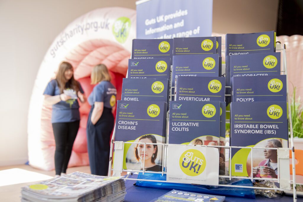 A selection of Guts UK patient information leaflets are displayed on a stand, on top of a table. Behind the stand, two people stand together talking.