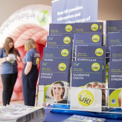 A selection of Guts UK patient information leaflets are displayed on a stand, on top of a table. Behind the stand, two people stand together talking.