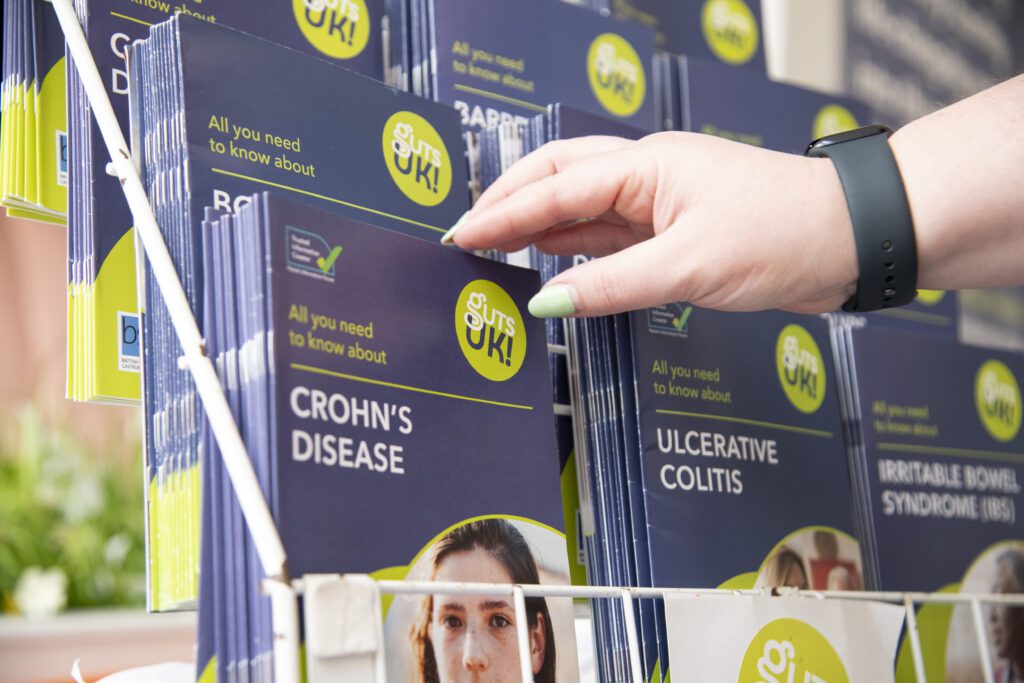 Guts UK's patient information leaflets are displayed in a stand. A hand reaches out to pick up the Crohn's disease leaflet.